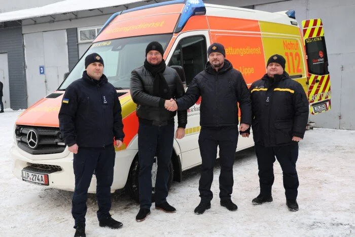 Чотири чоловіки біля помаранчево-білого рятувального мікроавтобуса тиснуть руки на снігу